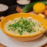 Cremiges Zitronen Risotto mit Zucchini, garniert mit frischer Petersilie. Zutaten wie Reis, Zitrone, Zucchini und Knoblauch sind im Hintergrund zu sehen.