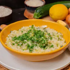 Cremiges Zitronen Risotto mit Zucchini, garniert mit frischer Petersilie. Zutaten wie Reis, Zitrone, Zucchini und Knoblauch sind im Hintergrund zu sehen.