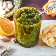 Ein Glas mit leuchtend grünem Pistazien Pesto, daneben eine Zitronenhälfte, einige Pistazien und eine Nudelrolle.