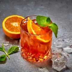 Ein erfrischender Cynar Spritz mit Orangenscheiben und Minze, umgeben von Eiswürfeln auf grauem Untergrund.