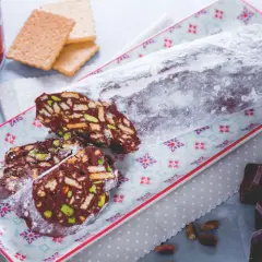 Ein köstlicher salame al cioccolato, bestäubt mit Puderzucker, auf einem bunten Teller. Daneben liegen zerbrochene Schokoladenstücke und Pistazien.