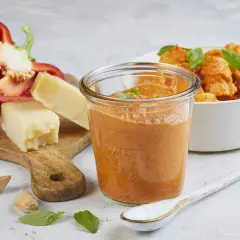 Ein Glas mit leuchtend rotem Paprika-Pesto steht neben einer Schüssel Pasta. Auf einem Holzbrett liegen eine halbe rote Paprika, Parmesankäse, Knoblauch und Basilikum.