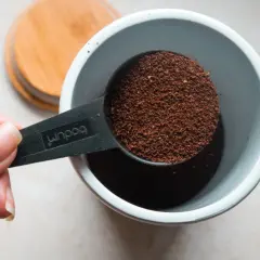Eine Hand hält einen Löffel mit Kaffeepulver, bereit, ihn in einen Behälter zu geben. Die Frage ist: wieviel kaffeepulver auf 1 liter wasser?