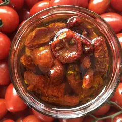 Ein Glas mit halbgetrockneten Tomaten, umgeben von frischen, roten Tomaten.