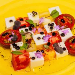 Tomaten-Mozzarella-Salat mit Balsamico-Kaviar, garniert mit Kräutern und Blüten auf gelbem Teller.