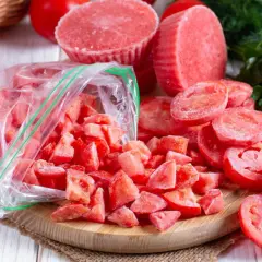 Gefrorene Tomatenstücke in einem Beutel und auf einem Holzbrett, bereit zum Einfrieren. Frische Tomaten und Petersilie im Hintergrund.