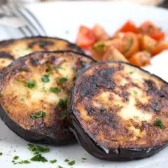 Drei Scheiben perfekt gebratene Aubergine, garniert mit Petersilie, serviert mit Kirschtomaten und einer cremigen Beilage.