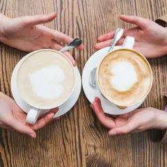 Zwei Hände reichen sich zwei Tassen mit cremigem Kaffee mit Milch, Latte Art in Herzform.