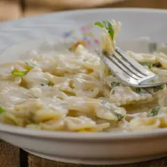 Farfalle-Nudeln in cremiger Sauce mit Zucchini und Ricotta, garniert mit frischen Kräutern. Ein Gabel hebt eine Portion an.