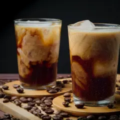 Zwei Gläser mit cremigem, kaltem Kaffee, umgeben von gerösteten Kaffeebohnen. Ein erfrischender Genuss an einem heißen Tag.