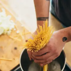 Hände mit Tattoos halten Spaghetti über einen Topf, um Nudeln zu kochen. Auf dem Holztisch liegt geschnittener Kohl.