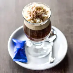 Ein Glas Kaffee mit Rum, gekrönt mit Sahne und Kakaopulver, serviert mit einer blauen Praline und Löffel auf einem Unterteller.