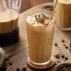 Cremiger Eiskaffee ohne Eis, garniert mit Sahne und Schokostreuseln, neben einer Kaffeemaschine und Kaffeebohnen.