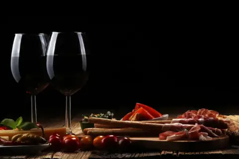 Zwei Gläser Rotwein, ein Cuvee Wein, stehen neben einer rustikalen Platte mit Käse, Schinken, Brot und Oliven.