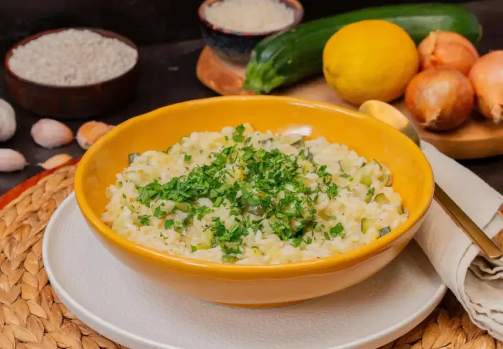 Cremiges Zitronen Risotto mit Zucchini, garniert mit frischer Petersilie. Zutaten wie Reis, Zitrone, Zucchini und Knoblauch sind im Hintergrund zu sehen.