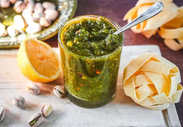 Ein Glas mit leuchtend grünem Pistazien Pesto, daneben eine Zitronenhälfte, einige Pistazien und eine Nudelrolle.