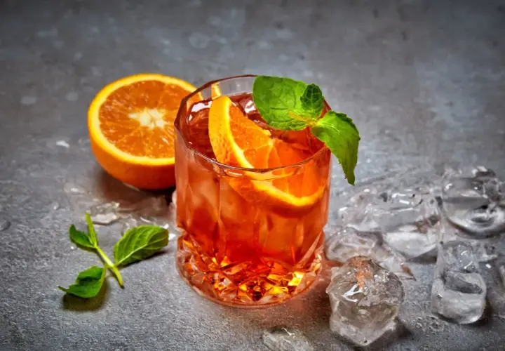 Ein erfrischender Cynar Spritz mit Orangenscheiben und Minze, umgeben von Eiswürfeln auf grauem Untergrund.