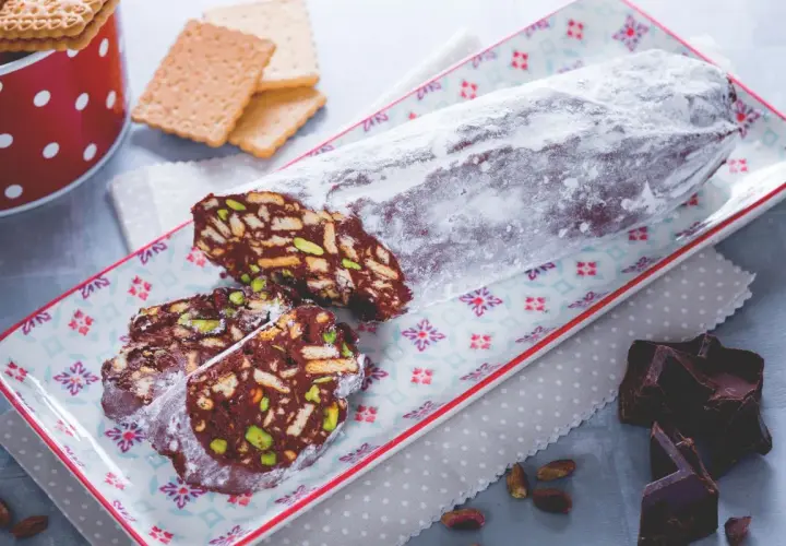 Ein köstlicher salame al cioccolato, bestäubt mit Puderzucker, auf einem bunten Teller. Daneben liegen zerbrochene Schokoladenstücke und Pistazien.