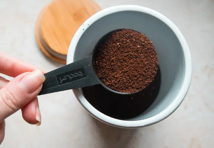Eine Hand hält einen Löffel mit Kaffeepulver, bereit, ihn in einen Behälter zu geben. Die Frage ist: wieviel kaffeepulver auf 1 liter wasser?