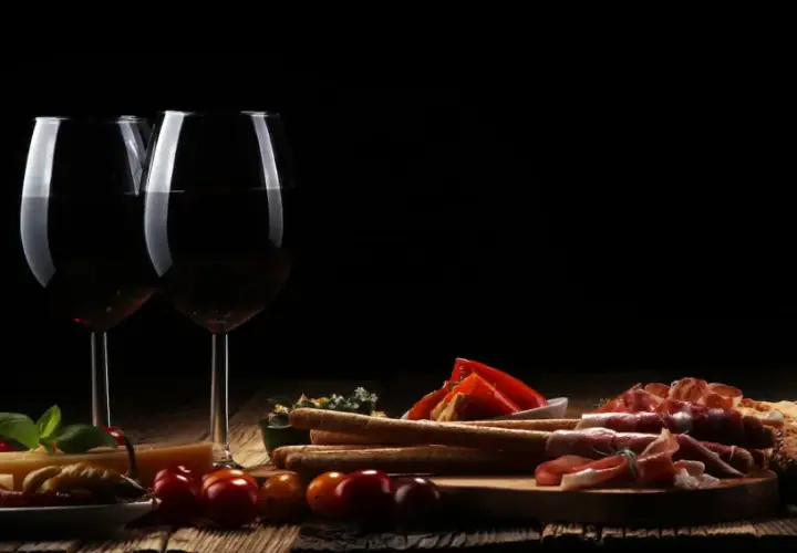 Zwei Gläser Rotwein, ein Cuvee Wein, stehen neben einer rustikalen Platte mit Käse, Schinken, Brot und Oliven.