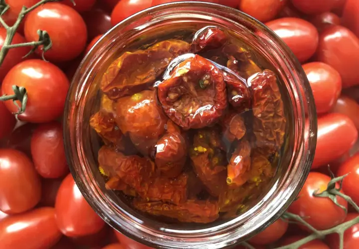 Ein Glas mit halbgetrockneten Tomaten, umgeben von frischen, roten Tomaten.