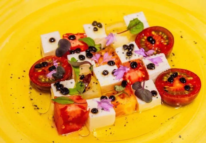 Tomaten-Mozzarella-Salat mit Balsamico-Kaviar, garniert mit Kräutern und Blüten auf gelbem Teller.