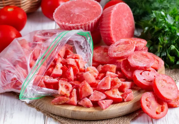 Gefrorene Tomatenstücke in einem Beutel und auf einem Holzbrett, bereit zum Einfrieren. Frische Tomaten und Petersilie im Hintergrund.