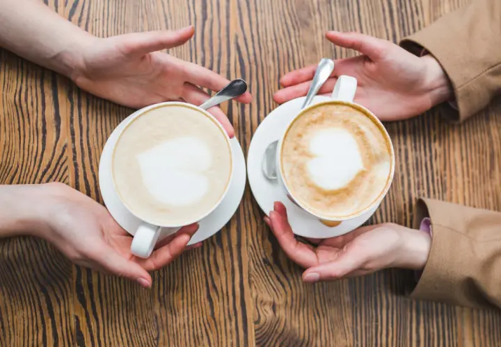 Zwei Hände reichen sich zwei Tassen mit cremigem Kaffee mit Milch, Latte Art in Herzform.