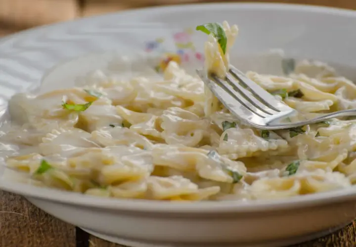 Farfalle-Nudeln in cremiger Sauce mit Zucchini und Ricotta, garniert mit frischen Kräutern. Ein Gabel hebt eine Portion an.