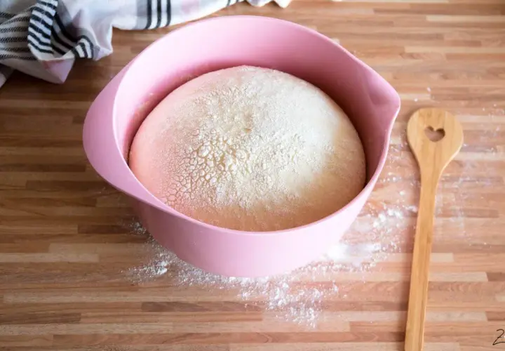 Ein aufgegangener Hefeteig in einer rosa Schüssel, bereit für die Zubereitung. Daneben liegt ein Holzlöffel mit Herz.