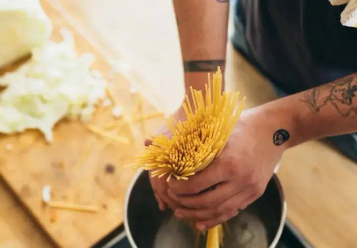 Hände mit Tattoos halten Spaghetti über einen Topf, um Nudeln zu kochen. Auf dem Holztisch liegt geschnittener Kohl.