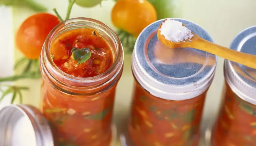 Frische Tomaten und Gläser mit Tomaten-Salsa, perfekt für leckere Tomaten Rezepte. Eine Löffelspitze Salz liegt bereit.