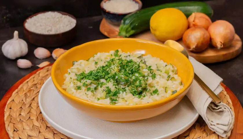 Cremiges Zitronen Risotto mit Zucchini, garniert mit frischer Petersilie. Zutaten wie Reis, Zitrone, Zucchini und Knoblauch sind im Hintergrund zu sehen.