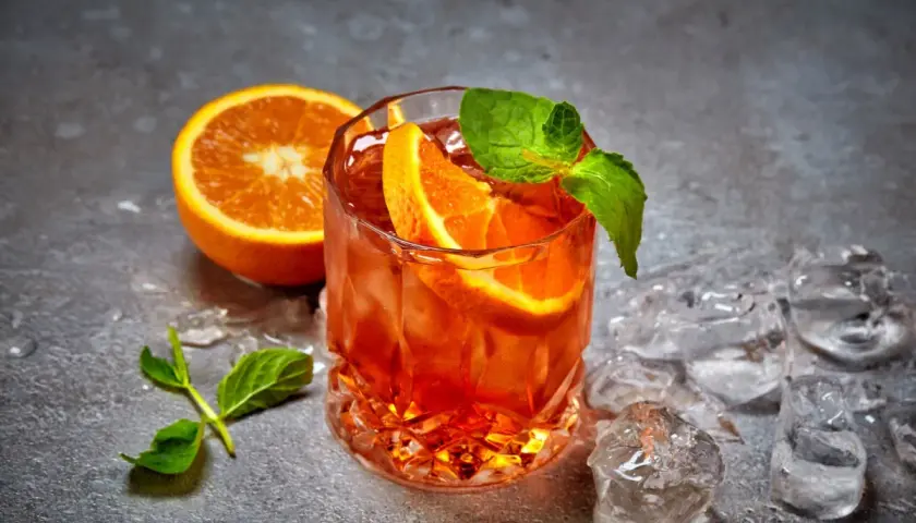 Ein erfrischender Cynar Spritz mit Orangenscheiben und Minze, umgeben von Eiswürfeln auf grauem Untergrund.