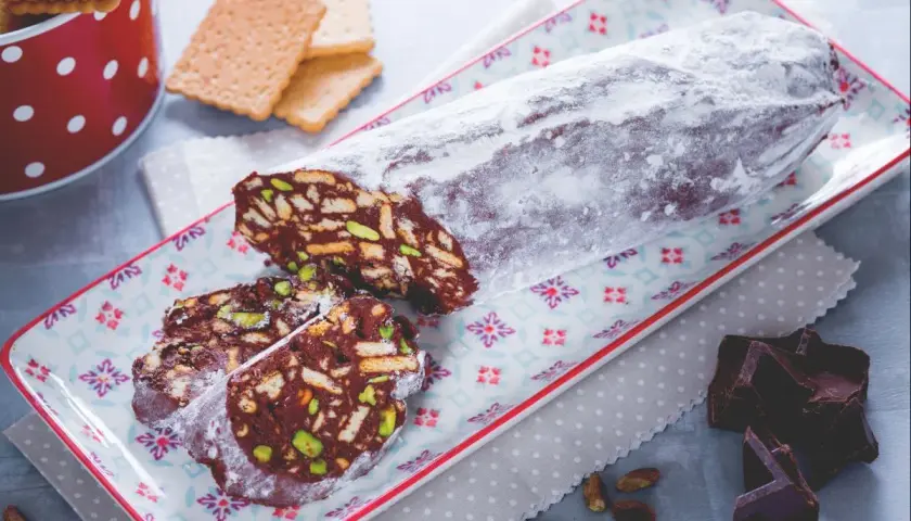 Ein köstlicher salame al cioccolato, bestäubt mit Puderzucker, auf einem bunten Teller. Daneben liegen zerbrochene Schokoladenstücke und Pistazien.