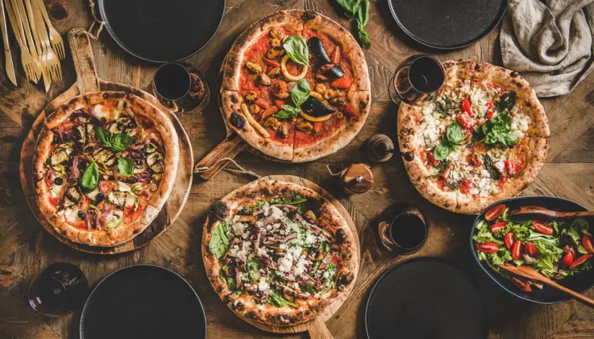 Vier leckere Pizzen, ein Salat und Rotwein auf rustikalem Holztisch. Perfekt für einen gemütlichen Abend mit Freunden.