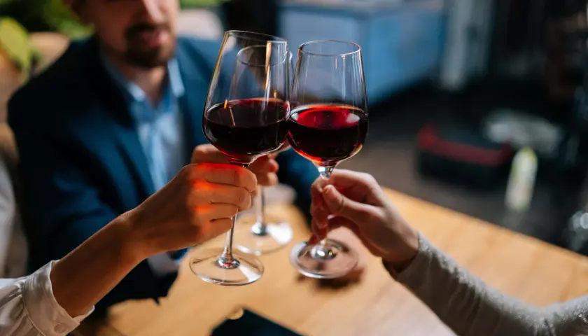 Drei Personen stoßen mit Rotwein an. Sie halten das weinglas richtig, am Stiel, um die Temperatur des Weins zu bewahren.