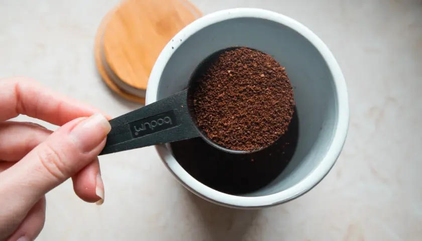 Eine Hand hält einen Löffel mit Kaffeepulver, bereit, ihn in einen Behälter zu geben. Die Frage ist: wieviel kaffeepulver auf 1 liter wasser?