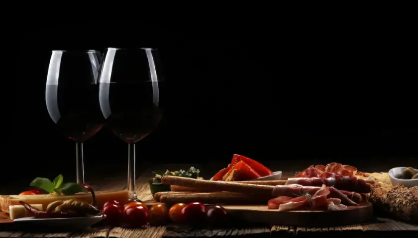 Zwei Gläser Rotwein, ein Cuvee Wein, stehen neben einer rustikalen Platte mit Käse, Schinken, Brot und Oliven.