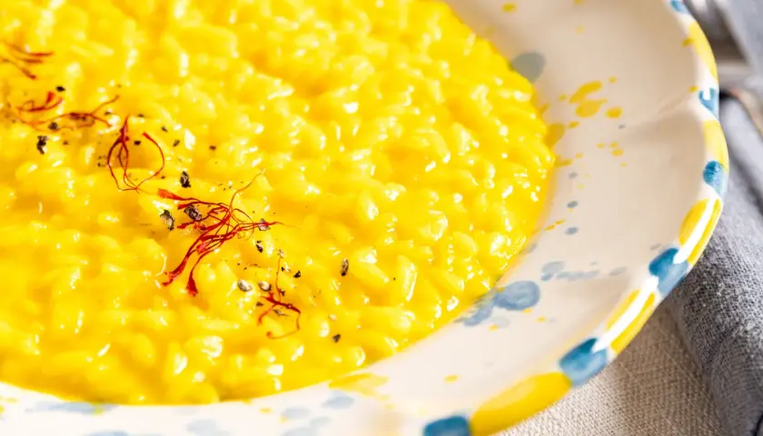 Cremiges, gelbes Risotto Milanese, garniert mit Safranfäden und schwarzem Pfeffer, in einem handbemalten Teller.