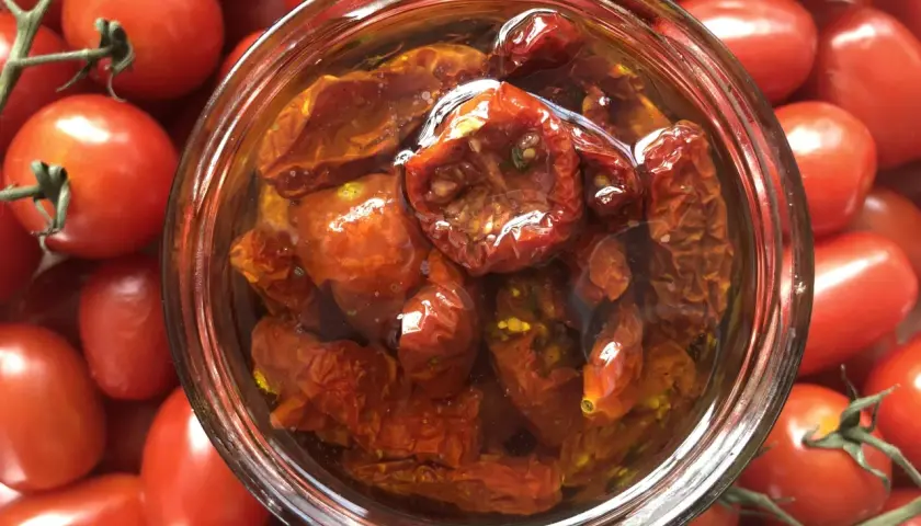 Ein Glas mit halbgetrockneten Tomaten, umgeben von frischen, roten Tomaten.