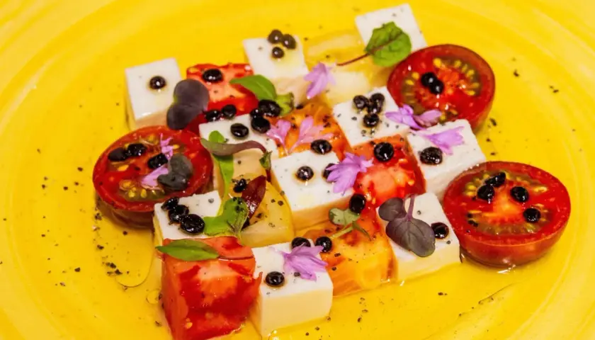 Tomaten-Mozzarella-Salat mit Balsamico-Kaviar, garniert mit Kräutern und Blüten auf gelbem Teller.