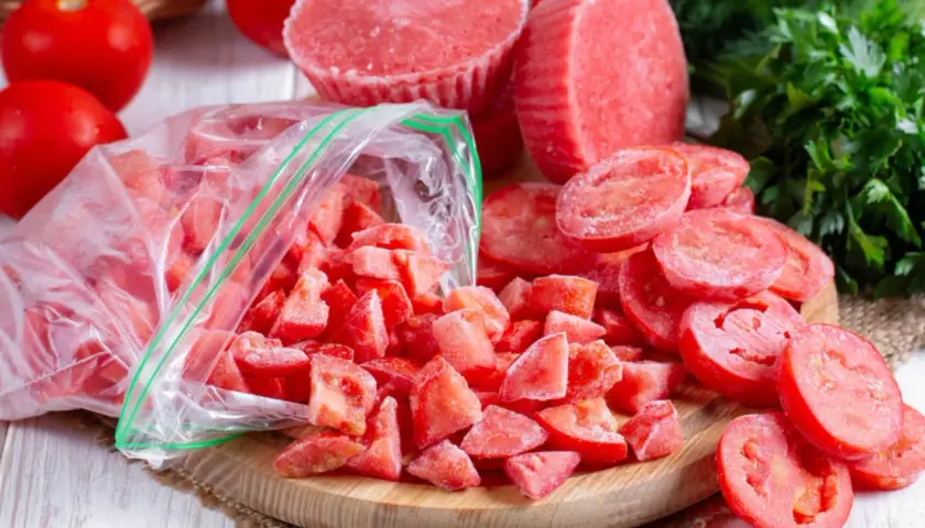 Gefrorene Tomatenstücke in einem Beutel und auf einem Holzbrett, bereit zum Einfrieren. Frische Tomaten und Petersilie im Hintergrund.