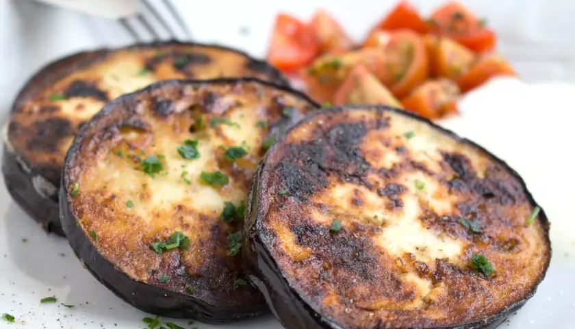 Drei Scheiben perfekt gebratene Aubergine, garniert mit Petersilie, serviert mit Kirschtomaten und einer cremigen Beilage.