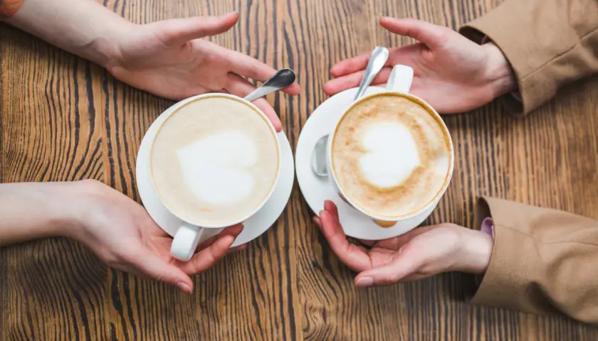 Zwei Hände reichen sich zwei Tassen mit cremigem Kaffee mit Milch, Latte Art in Herzform.