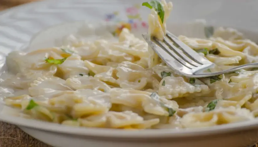 Farfalle-Nudeln in cremiger Sauce mit Zucchini und Ricotta, garniert mit frischen Kräutern. Ein Gabel hebt eine Portion an.