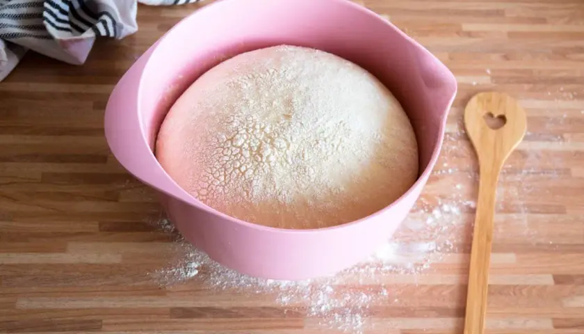 Ein aufgegangener Hefeteig in einer rosa Schüssel, bereit für die Zubereitung. Daneben liegt ein Holzlöffel mit Herz.