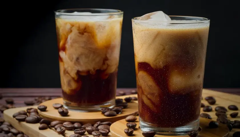 Zwei Gläser mit cremigem, kaltem Kaffee, umgeben von gerösteten Kaffeebohnen. Ein erfrischender Genuss an einem heißen Tag.