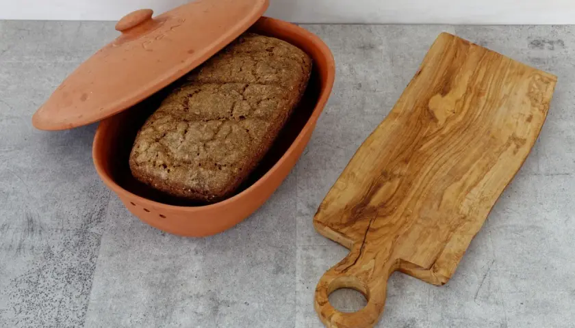 Ein Laib Brot liegt in einem Terrakotta-Brotbehälter. Daneben eine Holzschneidebrett. Perfekt, um Brot aufbewahren.