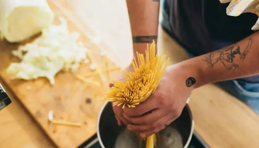 Hände mit Tattoos halten Spaghetti über einen Topf, um Nudeln zu kochen. Auf dem Holztisch liegt geschnittener Kohl.